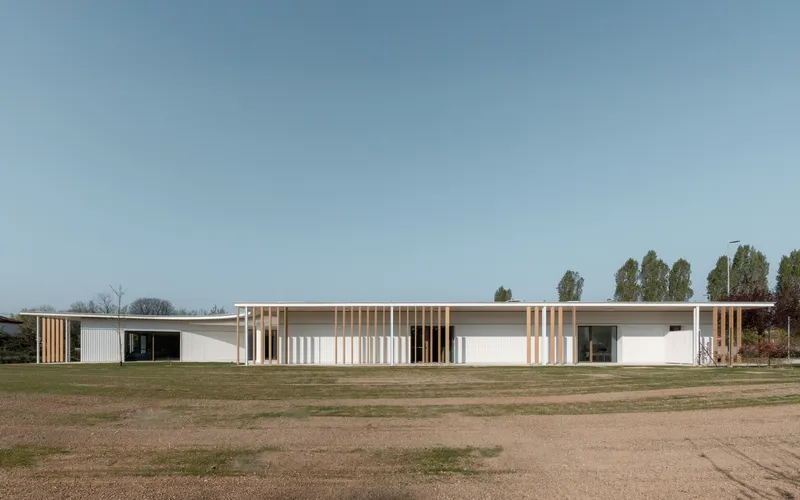 Long facade showing alternating white panels and timber screens facing an open field with distant trees