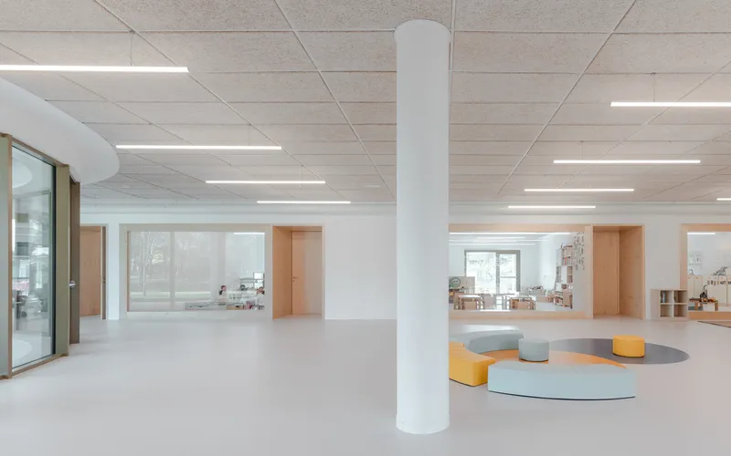 Interior space with white ceiling panels, linear lighting, and circular seating elements on pale flooring