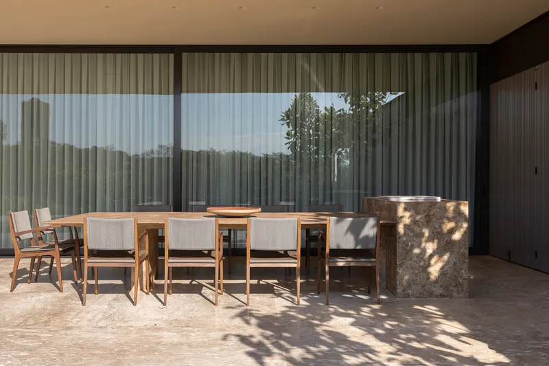 Covered terrace with dining table and chairs casting dappled shadows under tree canopy beyond glass