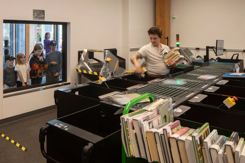 Automated book sorting conveyor belt with green bins and onlookers visible through a glass window