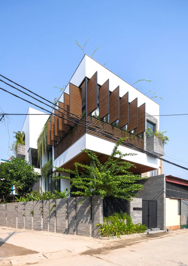 Corner facade featuring angled timber louvers shading upper balconies above a concrete boundary wall with trees