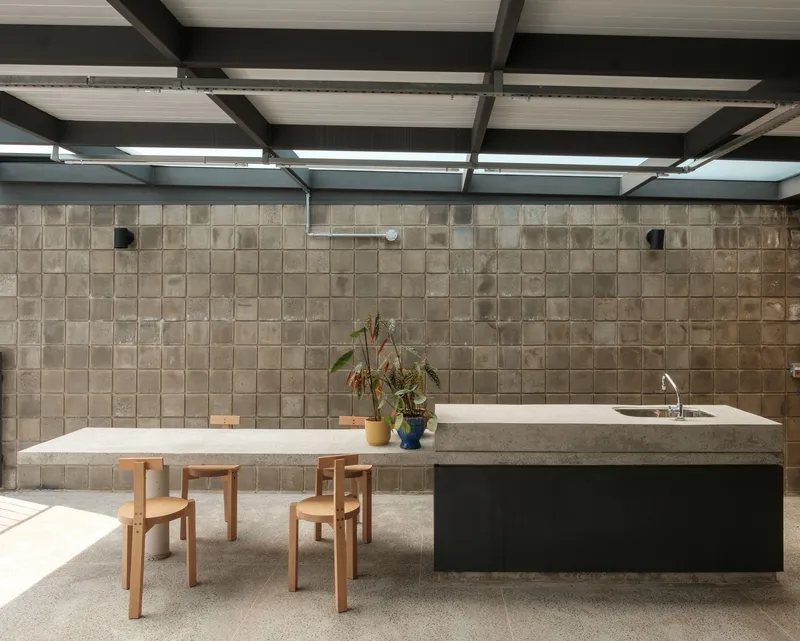 Dining area with concrete island, timber chairs, and beige tile wall under exposed steel ceiling beams