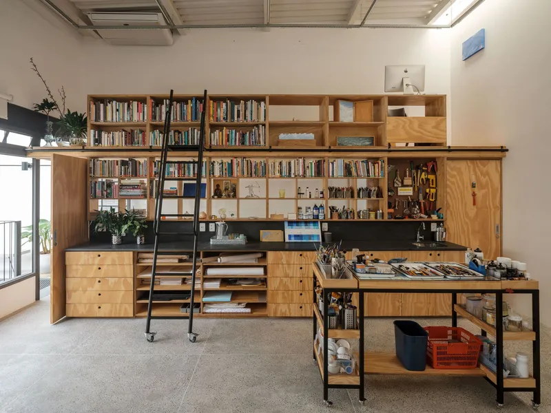 Floor-to-ceiling plywood storage wall with rolling ladder and black steel work table in foreground
