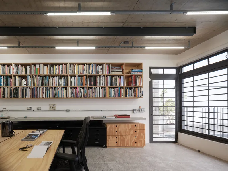 Wall-mounted timber bookshelves above built-in desk with gridded window screen and exposed concrete beams