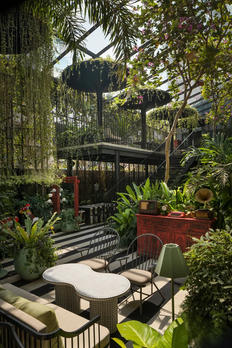 Outdoor seating area beneath a planted steel trellis system with hanging vines and tropical foliage