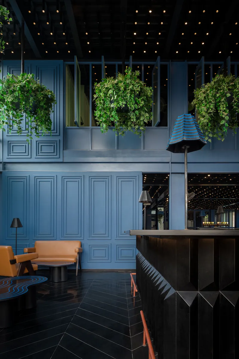 Double-height interior with blue paneled walls, suspended planters, and herringbone tile flooring at night