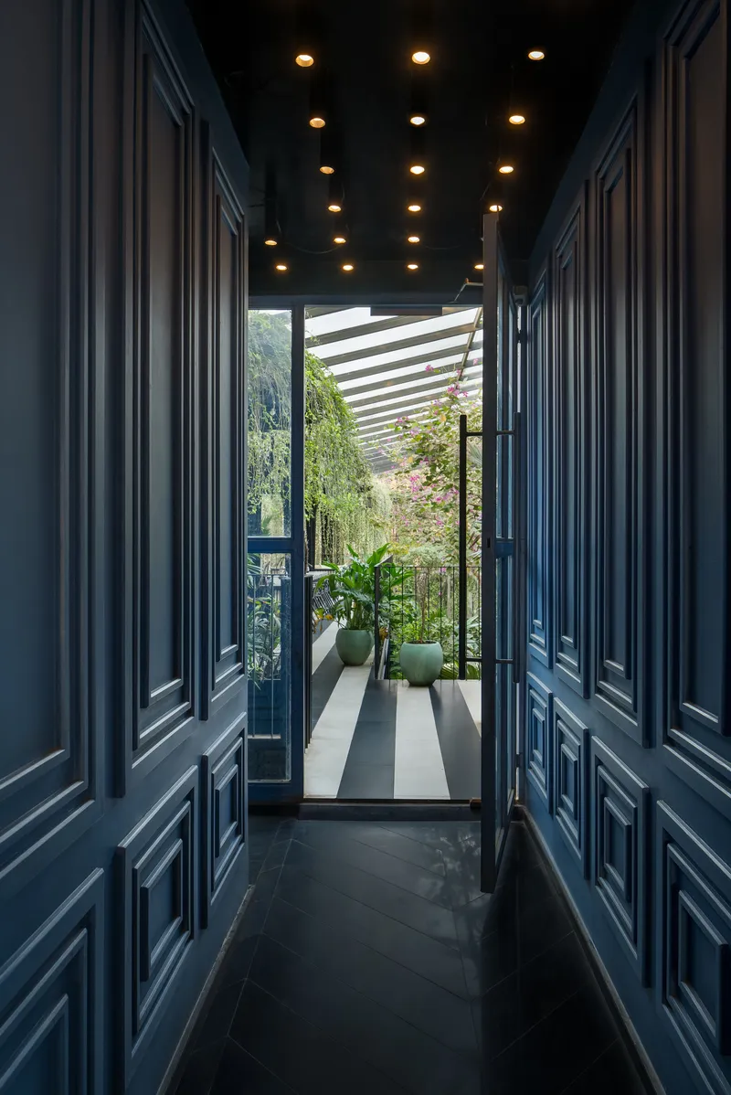 Narrow corridor with navy paneled walls and ceiling-mounted downlights leading to a planted terrace with slatted pergola