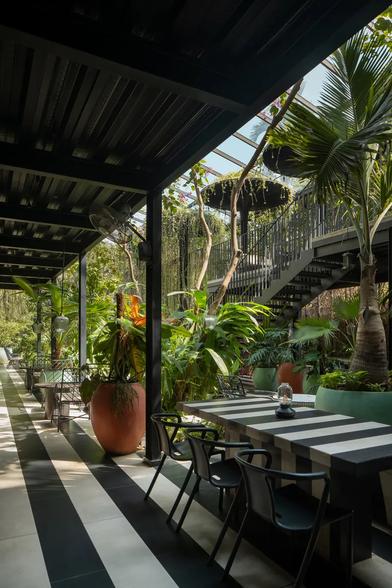 Covered dining terrace with exposed timber beams and striped floor tiles adjacent to garden with palms and hanging plants