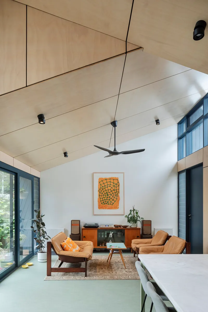 This architectural image showcases a modern living room with a vaulted plywood ceiling and abundant natural light. The space features mid-century modern furnitu