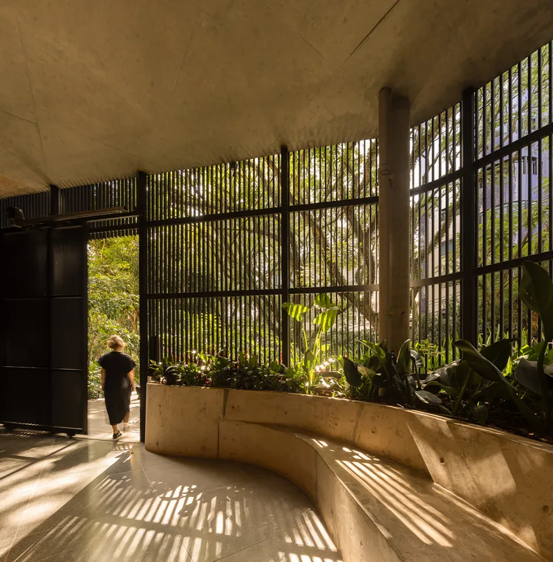 Lobby space with vertical brise-soleil casting striped shadows across curved concrete planter and stone floor