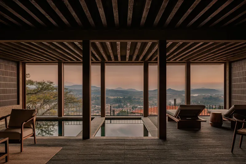 Interior pavilion with exposed timber ceiling beams and floor-to-ceiling glazing framing distant mountains at dusk