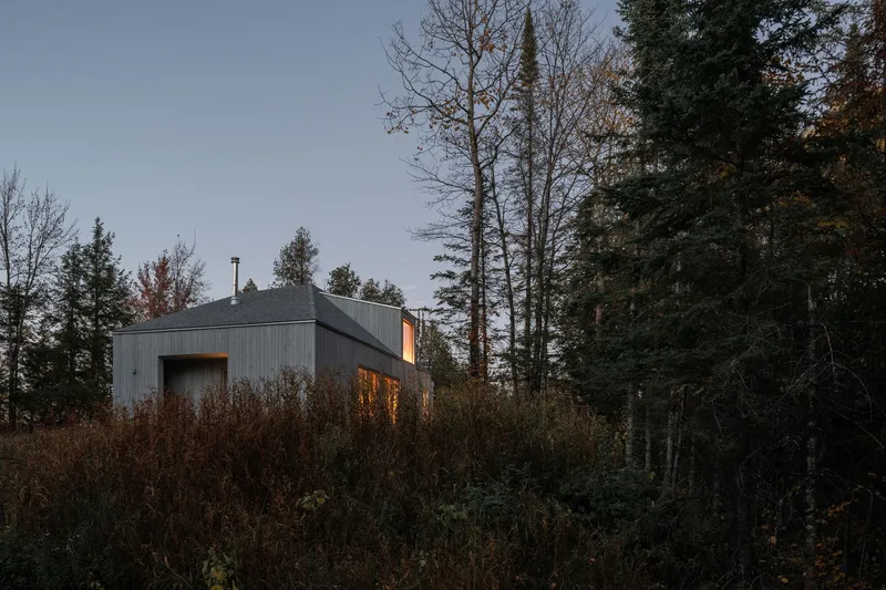 Grey corrugated metal clad volume nestled among bare deciduous and evergreen trees at dusk