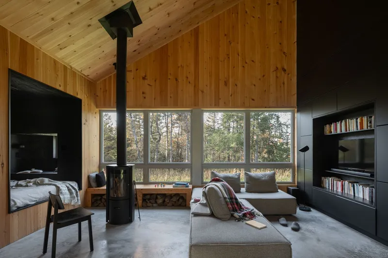 Living area with freestanding wood stove and horizontal windows framing meadow and trees beyond