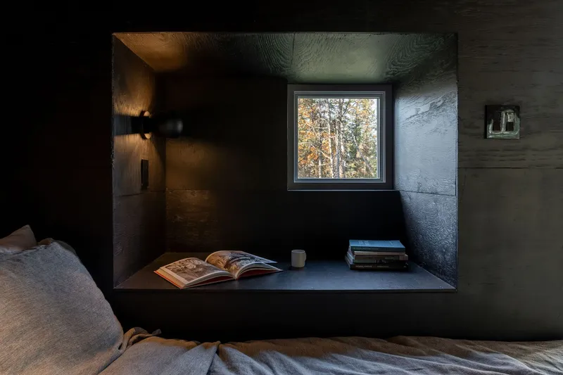 Built-in shelf with square window framing trees in blackened plywood bedroom alcove