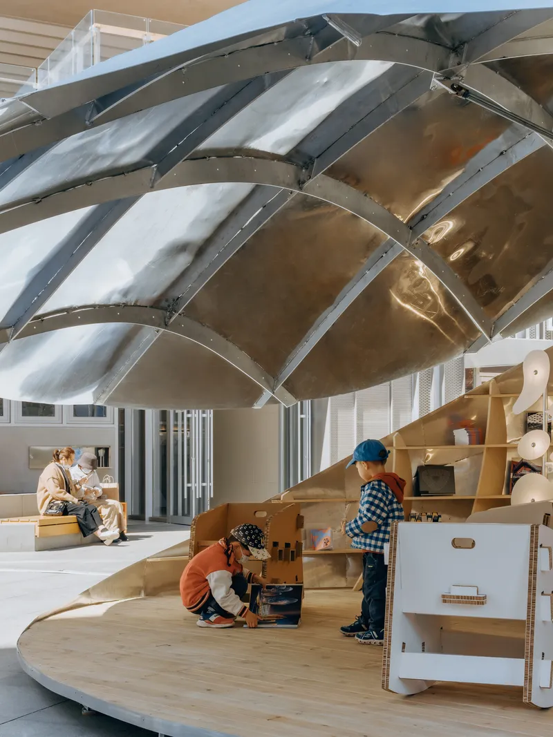Interior view of the ribbed metal shell structure above children playing with modular cardboard toys