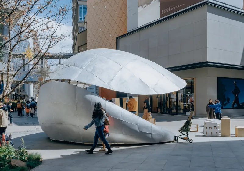 Shell-shaped pavilion with white fabric canopy and grey base on a plaza with pedestrians and bare trees