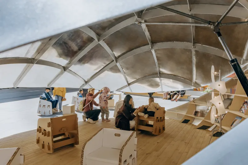 Interior view under the ribbed translucent canopy with children assembling cardboard furniture on the plywood floor