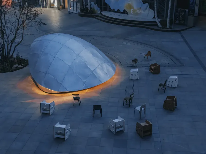 Aerial view of illuminated pod structure surrounded by scattered chairs and cardboard furniture at dusk