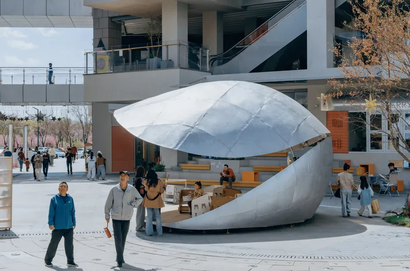 Shell pavilion on public plaza with visitors and autumn trees in front of glass and concrete building