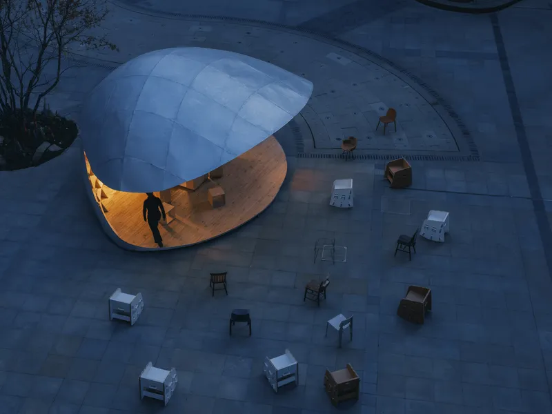 Aerial view of the illuminated pavilion at dusk surrounded by scattered furniture on paved plaza
