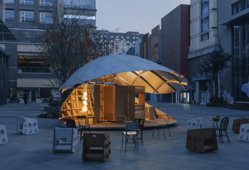 Timber shell pavilion with ribbed canopy and glowing interior in an urban plaza at dusk