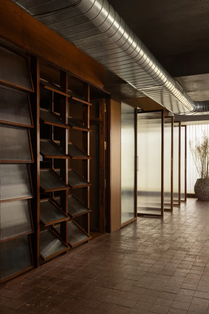 Corridor with exposed ductwork ceiling and geometric wooden shelving beside translucent glass partitions