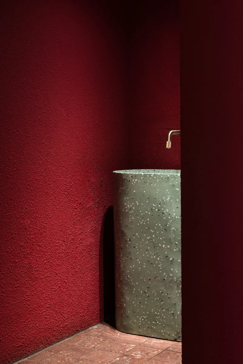 Deep red plaster wall corner with terrazzo pedestal sink and brass wall-mounted tap