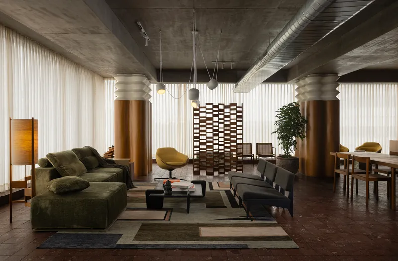 Open living space with exposed concrete ceiling, cylindrical bronze columns, and sheer curtains filtering daylight