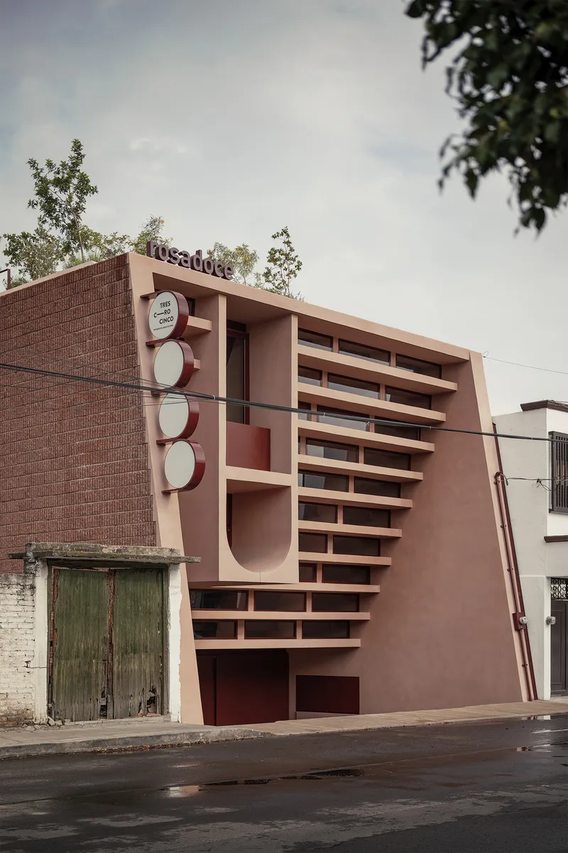 Street view of the pink stucco facade with horizontal louvers and circular motifs beside a brick wall