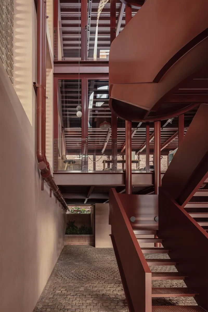Narrow courtyard passage with red steel stairs and bridges connecting levels beneath slatted screens