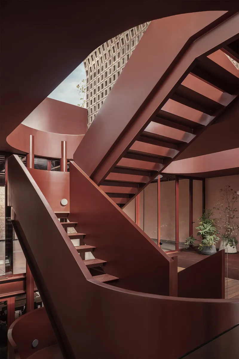 Red steel staircase with open risers ascending through a skylight framing a perforated brick facade