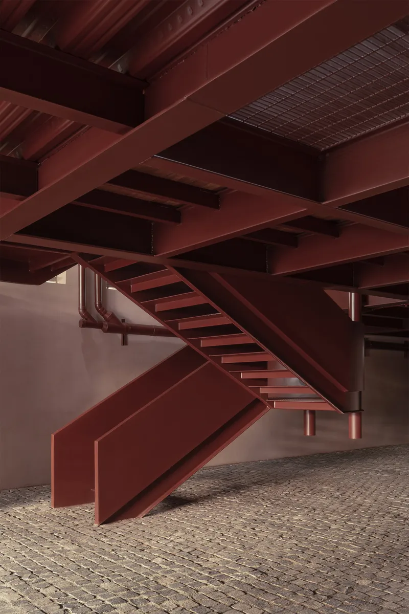 Underside view of intersecting steel beams and staircases with exposed structural grid above cobblestones