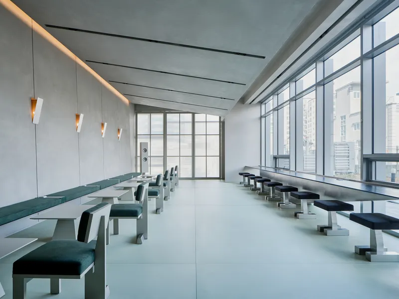 Communal room with paired tables and seating along glazed walls under a ceiling with recessed linear lights