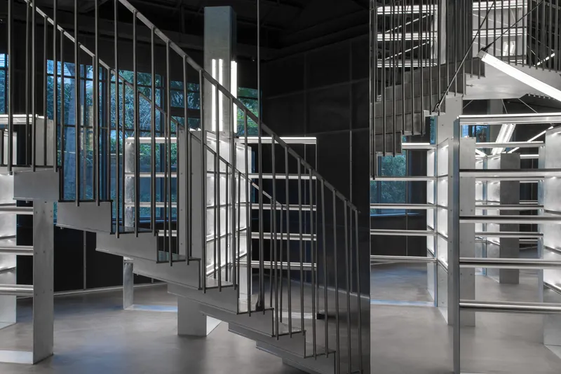 Open staircase with vertical metal railings connecting multiple levels under a black paneled ceiling with daylight filtering through