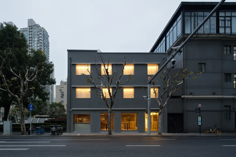 Three-storey grey facade with illuminated windows and bare street trees at dusk