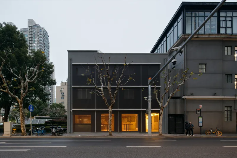 Street view of the grey box facade with pedestrians gathered near bare trees at twilight