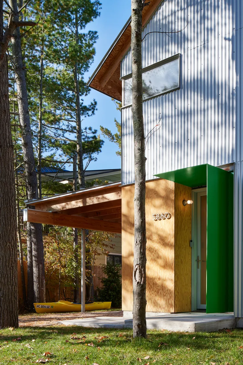Entry canopy with timber roof and green metal column among pine tree trunks casting shadows