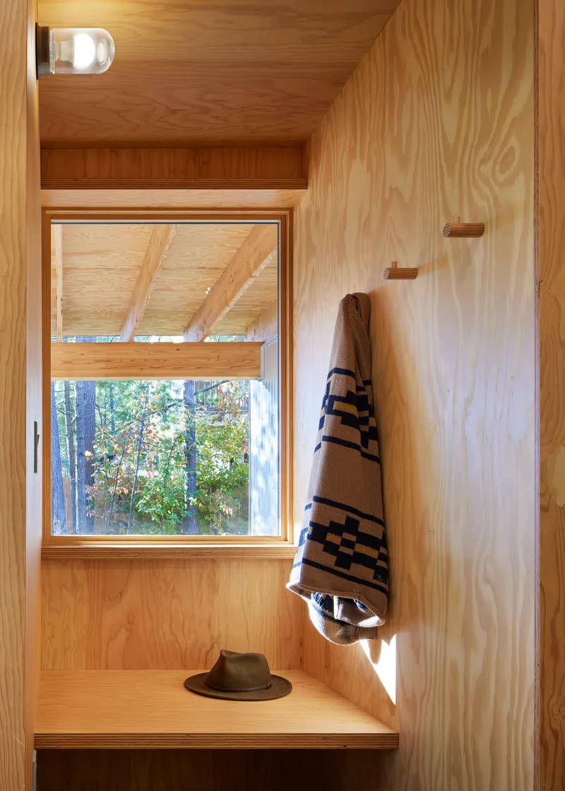 Plywood-lined entryway nook with bench, wall hooks, and window overlooking forest in afternoon light