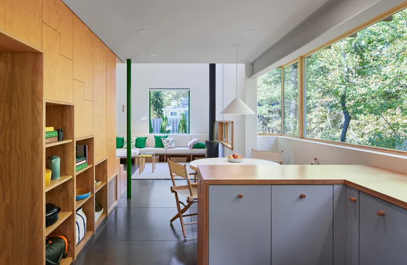 Open kitchen and living area with plywood shelving, grey island, and full-height windows facing trees