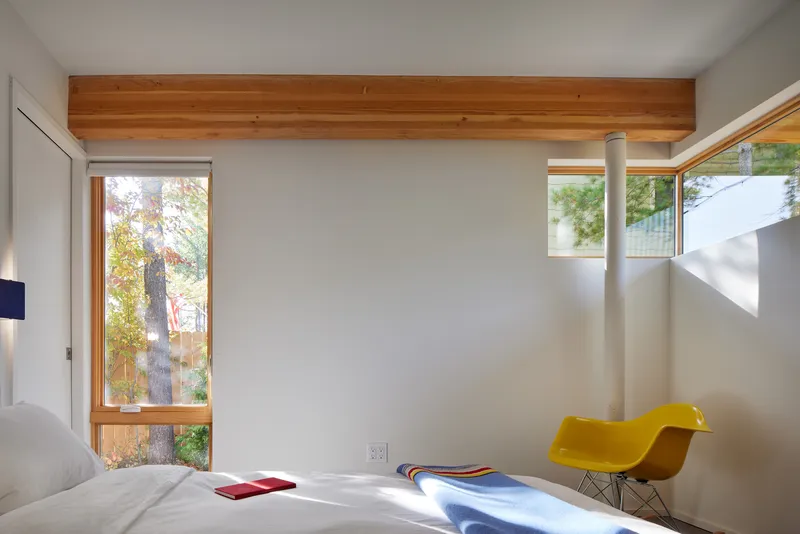 Bedroom with timber ceiling beam, tall window framing birch trees, and yellow chair catching sunlight