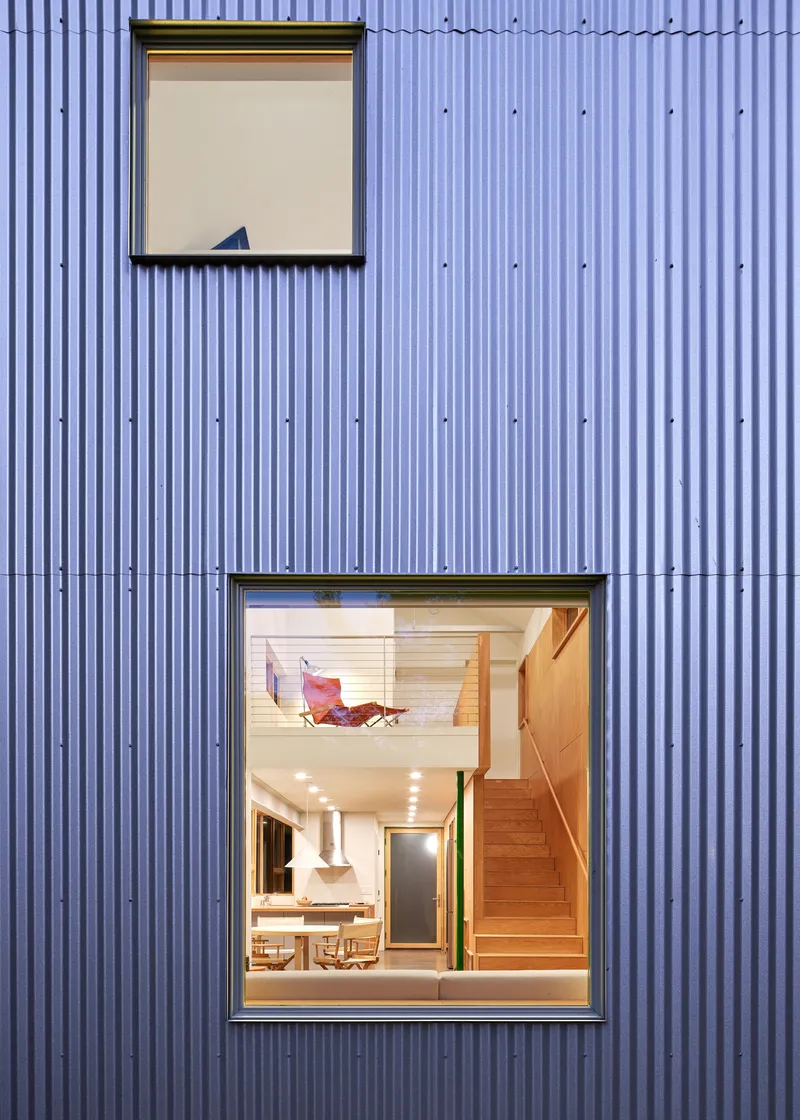 Facade detail of corrugated metal siding with two stacked windows revealing plywood interior and staircase at dusk
