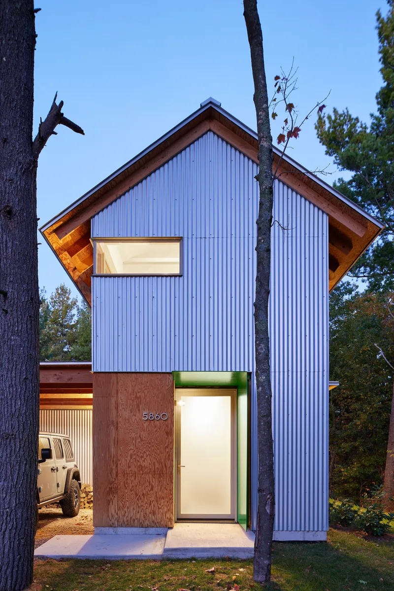 Vertical corrugated metal facade with recessed entry and carport framed by pine trees at dusk