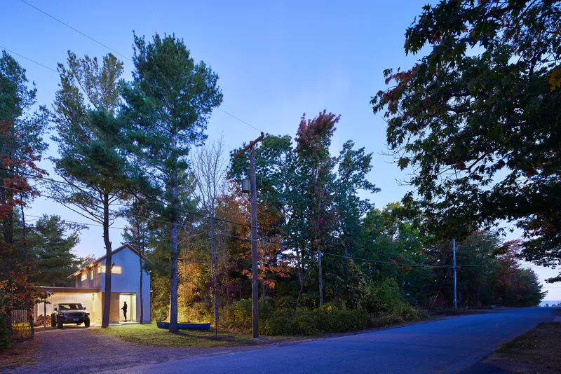 Street view of the gabled volume nestled among trees with illuminated windows at twilight