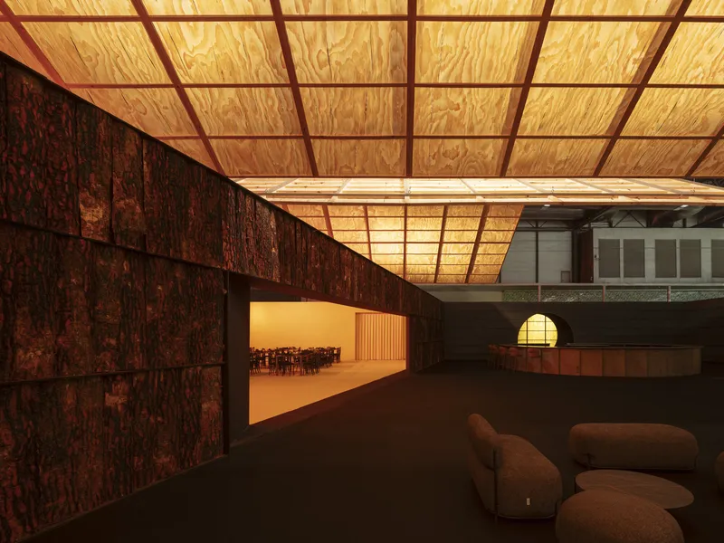 Interior space with plywood panel ceiling grid and textured cork wall beside a sunlit corridor