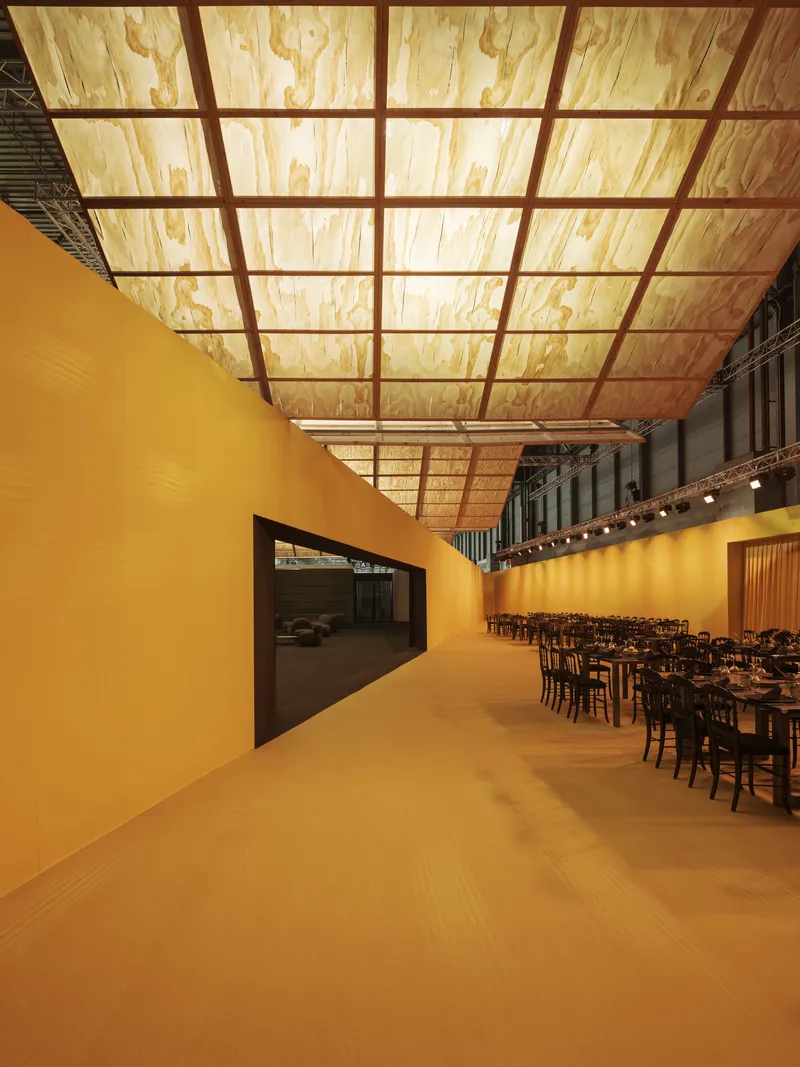 Corridor with ochre walls and backlit plywood ceiling grid leading to a dining area