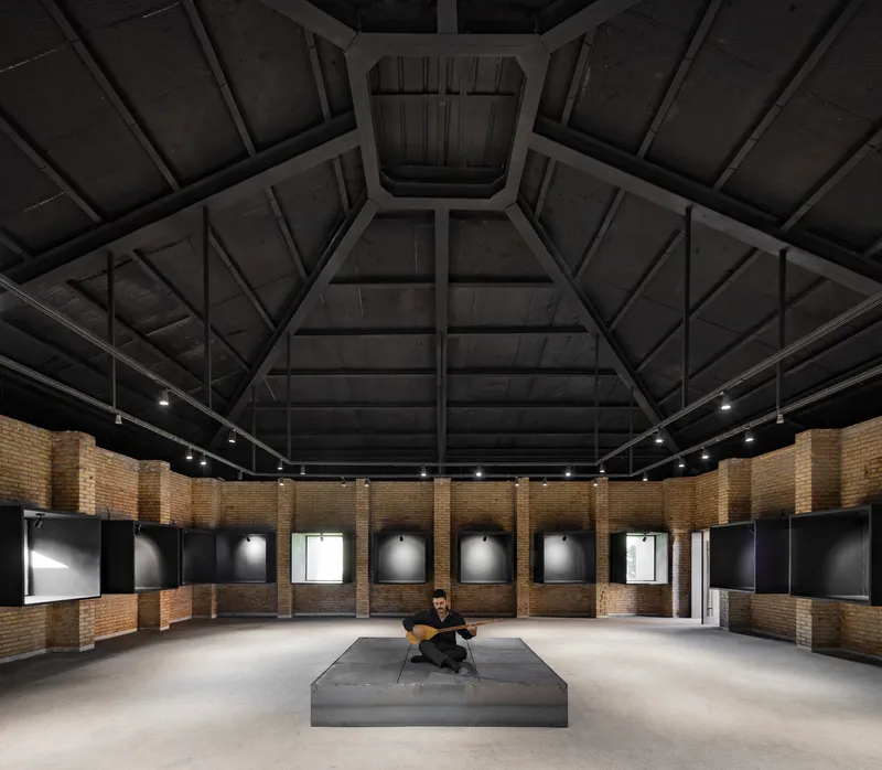 Interior gallery space with exposed timber roof beams radiating from a central skylight above a seated figure