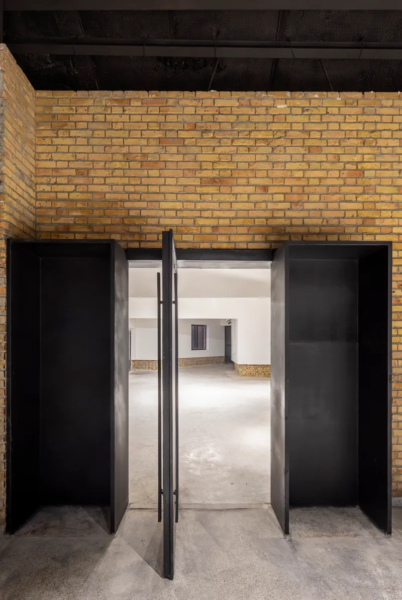 Pivot doors in black metal frames opening from a yellow brick wall into white-walled interior spaces