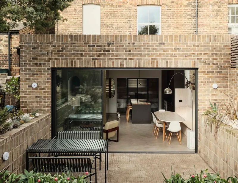 Rear courtyard with full-height glass doors opening to a kitchen and dining area beneath buff brick walls