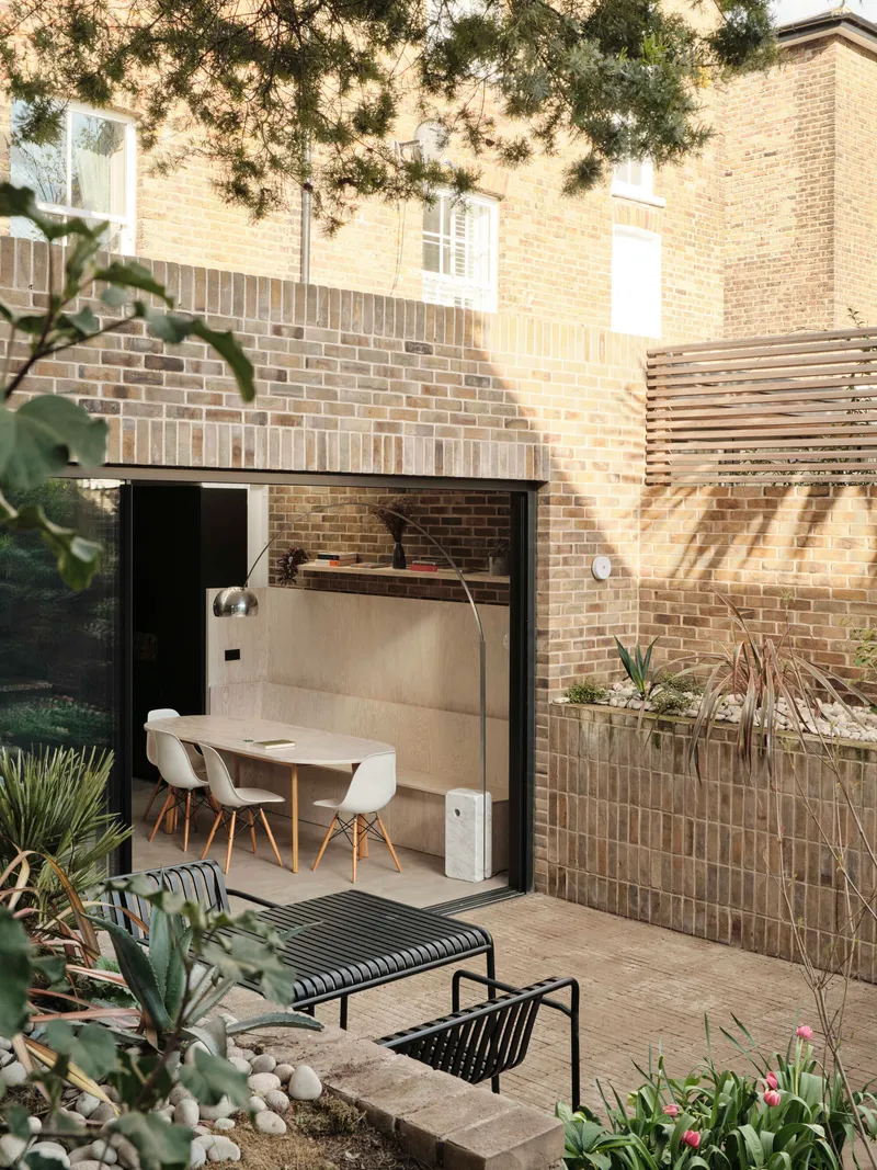 Glazed rear extension opening to courtyard garden with lounge chairs beneath overhanging tree branches