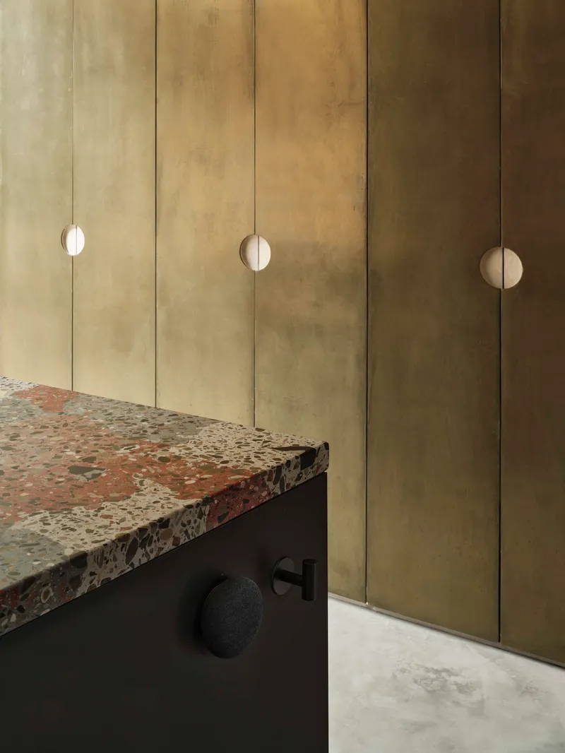 Brushed brass cabinet wall with circular recessed pulls beside a dark joinery unit with terrazzo countertop